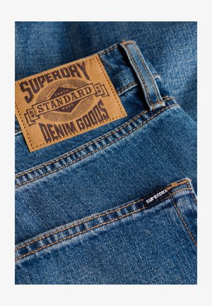Blauwe denimjeans met een bruine leren patch met het label "SUPERDRY STANDARD DENIM GOODS" en een klein zwart label op de zak.