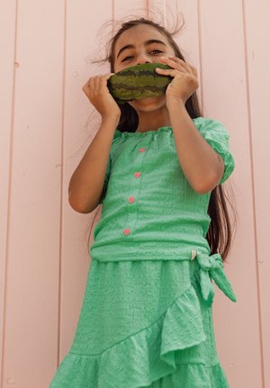 Jong meisje in een groen jurkje bijt speels in een kleine watermeloen tegen een lichtroze houten muur als achtergrond.