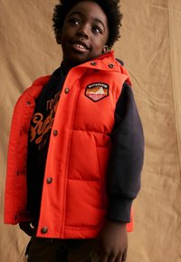 Gilet rembourré orange vif avec des boutons et des poches latérales, doté d'une manche noire et d'un patch logo sur la poitrine, porté par-dessus un hoodie sombre.