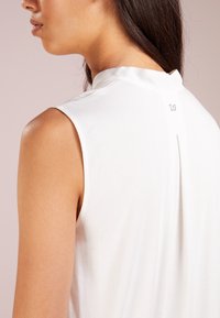 Top sin mangas blanco con un cuello alto, textura suave y un pliegue en la parte posterior, acentuado por un pequeño detalle de logo cerca del escote.