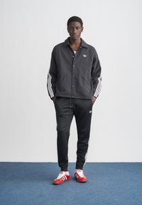 Črna jakna na gumbke z belo Adidasovo logotipom in rokavi s tremi črtami, kombinirana s črnimi športnimi hlačami in rdečimi supergami.