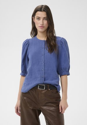 Ženska z dolgimi rjavimi lasmi, oblečena v modro bluzo z gumbi in napihnjenimi rokavi ter rjave usnjene hlače s črnim tkanim pasom.