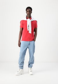 Polo rouge, jean bleu clair, baskets blanches et un sweat-shirt gris drapé sur les épaules. Tissus lisses, coupe décontractée et couleurs contrastées.
