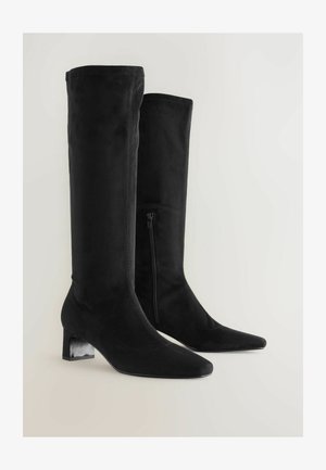 Botas de ante negras hasta la rodilla con punteras puntiagudas, acabado texturizado y tacón cuadrado. Cierre lateral para facilitar el uso.