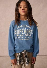 Felpa blu con testo bianco "SUPERDRY" e logo. Abbinata a pantaloni corti kaki e una camicia a quadri legata in vita. Tessuto testurizzato.