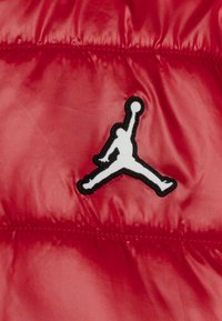 Punainen puffatakkia, jossa on kiiltävä pinta, ja edessä musta-valkoinen Jumpman-logo-ompelus.