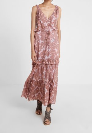 Kvinde iført en ærmeløs, lyserød mønstret maxikjole med flæser og brune åbne hælede sandaler, stående på et hvidt gulv.