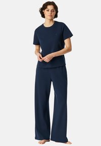 Das marineblaue Loungewear-Set umfasst ein Kurzarmshirt und weite Hose, gefertigt aus einem weichen Stoff mit schlichtem Design und dezentem Logo-Detail.