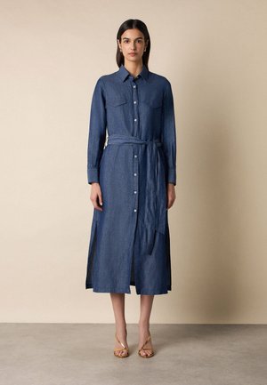 Kobieta stoi przy beżowej ścianie, ubrana w niebieską midi sukienkę z długim rękawem i guzikiem, z paskiem w talii, oraz złote sandały na obcasie na szarej podłodze.