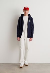 Mørkeblå hettejakke med glidelås over en hvit t-skjorte, kombinert med hvite bukser og hvite joggesko. Modellen har også på seg en rød baseballcap.