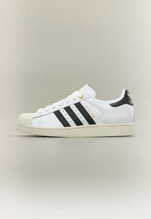 Zapatilla Adidas blanca de corte bajo con rayas negras, puntera texturizada y letras doradas "Superstar" en el lateral.