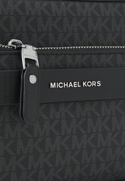 Sac à main en tissu noir présentant un motif texturé, des accessoires en argent, et une bandoulière en cuir noir distincte avec un logo en relief.