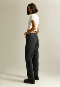 Mujer con cabello corto castaño, vistiendo una blusa blanca ajustada, jeans negros holgados y zapatos negros, de pie frente a un fondo beige liso.