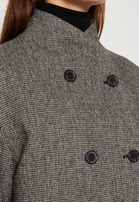 Femme portant un manteau à motif pied-de-poule à double boutonnage avec des boutons noirs par-dessus un pull col roulé noir.