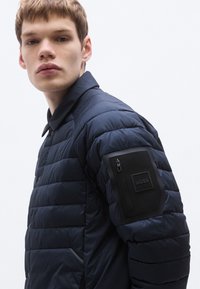 Granatowa kurtka puchowa z pikowaną fakturą, stójką oraz zapinaną na zamek kieszenią na lewym ramieniu, na której znajduje się czarna naszywka z logo.