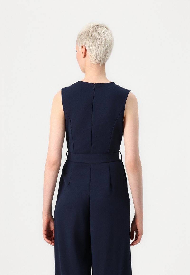 Tuta blu navy con un corpetto strutturato, maniche a cappuccio e vita definita. Presenta pieghe sul retro e una cerniera nascosta. Tessuto liscio.