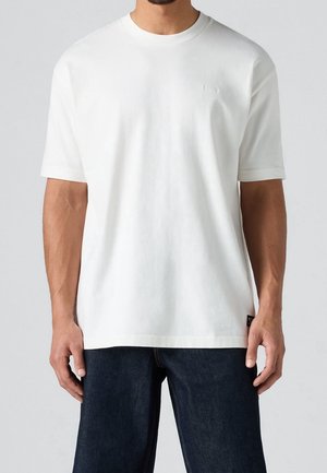 Homme portant un T-shirt blanc uni à manches courtes et un jean foncé, vu des épaules aux cuisses, debout devant un fond gris clair.