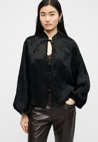 Γυναίκα που φοράει μαύρο σατέν μπλουζάκι με μπαλόν μανίκια και κλείσιμο με κορδόνια πάνω από δαντελένιο τοπ, συνδυασμένο με παντελόνι από σκουρό καφέ δέρμα.