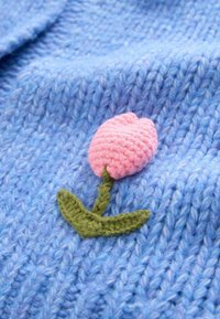 Světlomodrá pletenina s růžovým háčkovaným tulipánem a zelenými listy, zobrazující texturu a živé barvy.