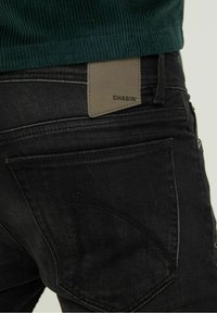 Jeans de mezclilla negra con una vista en primer plano del bolsillo trasero y la etiqueta de cuero con la marca "CHASIN". Tejido texturizado con detalles de costura visibles.