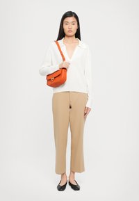 Woman in a white V-neck sweater, beige cropped trousers, black flats, holding an orange shoulder bag against a plain white background.