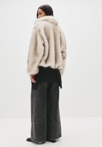 Jasno beżowa kurtka z futra sztucznego z szerokim kołnierzem, zestawiona z czarnym, warstwowym topem i ciemnymi, szerokimi dżinsami z postrzępionymi nogawkami.