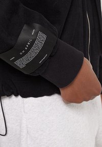 Czarna bluza z kapturem na zamek, z prążkowanymi mankietami, z falistą metką na lewym rękawie z napisem. Nosi się ją na jasnoszarych spodniach.