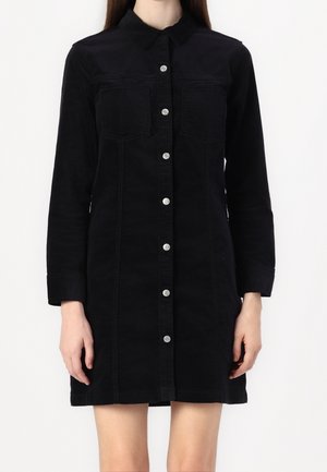 Femme portant une robe noire à manches longues boutonnée avec des poches poitrine, debout devant un fond blanc uni.