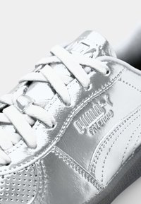 Baskets métalliques argentées avec une finition lisse et brillante, des lacets blancs, un panneau texturé et un logo Puma embossé sur le côté.