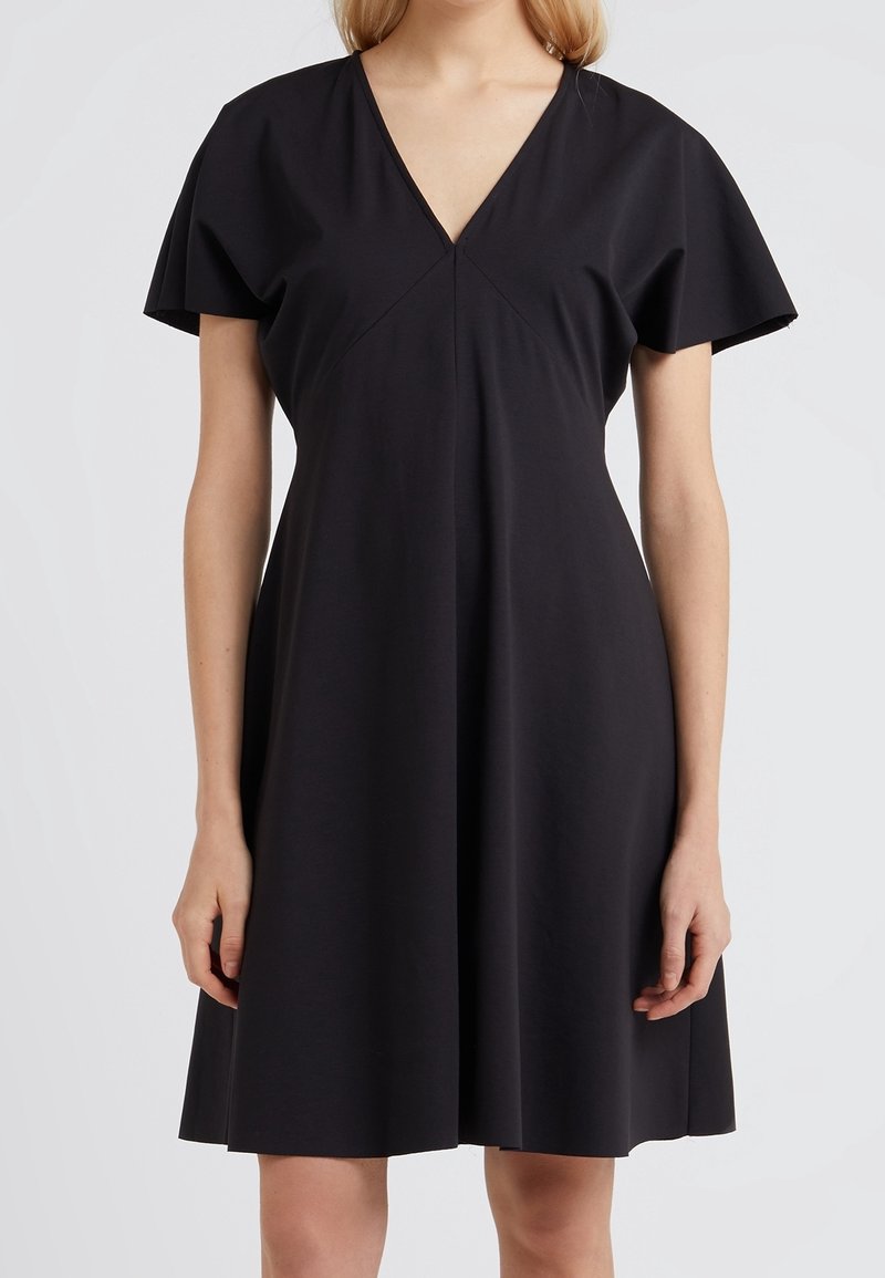Femme portant une robe noire à manches courtes et décolleté en V, avec une silhouette A-line ample, sur un fond clair uni.
