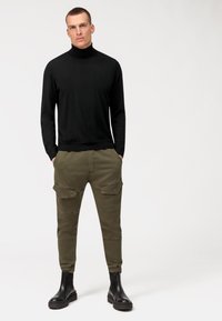 Schwarzer Rollkragenpullover, olivgrüne Cargo-Hose mit Seitentaschen und schwarze Stiefeletten. Glatte Textur und figurbetontes Design, das den lässigen Stil betont.