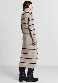 Abito lavorato a maglia beige con strisce orizzontali nere, colletto alto, maniche lunghe e spacchi laterali, abbinato a stivali neri al ginocchio.