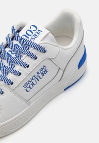 White sneaker with blue accents, textured leather, bold logo, and blue and white striped laces, featuring a padded ankle collar.