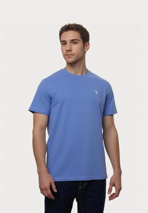 Junger Mann, der ein schlichtes hellblaues T-Shirt mit kurzen Ärmeln und eine dunkle Hose trägt, steht vor einem einfarbigen weißen Hintergrund.