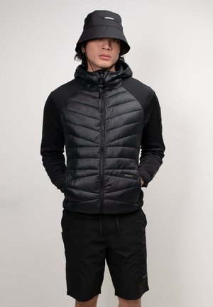 Junge Person in komplett schwarzem Outfit mit einem Fischerhut, gesteppter Reißverschlussjacke und Shorts, steht mit den Händen in den Taschen vor einem einfarbigen Hintergrund.