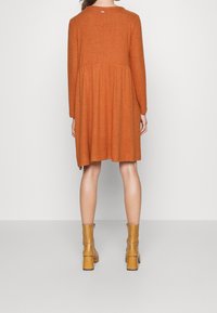 Vestido de manga larga en color óxido con un diseño acampanado y escote redondeado. Textura suave con una costura en la cintura. Combinado con botas de tobillo en tonos beige.