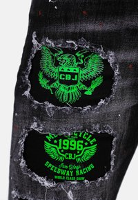 Jeans in denim nero con effetti vissuti e ampie aperture che rivelano toppe grafiche verdi. Il design presenta loghi a tema motociclistico e dettagli a forma di scudo.