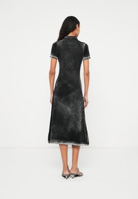 Femeie cu păr lung și creț, purtând o rochie midi neagră cu mâneci scurte spălată, și sandale argintii cu toc, stând cu fața spre spate.