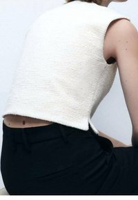 Biała, teksturowana bezrękawnikowa bluzka o skróconym kroju, zestawiona z czarnymi, eleganckimi spodniami. Posiada boczne rozcięcia przy dolnym brzegu dla dodatkowego efektu.