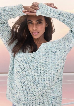 Hellblauer Strickpullover mit V-Ausschnitt und gerippten Bündchen, der ein strukturiertes Muster und dezente grüne Akzente aufweist. Modell posiert mit erhobenen Armen.