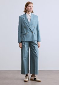 Lichtblauw, double-breasted pak met gouden knopen, wijde broekspijpen, gecombineerd met een wit overhemd en casual sneakers. Soepel stoftextuur.