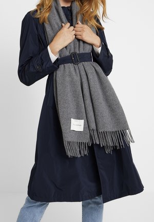 Personne portant un trench-coat bleu marine ceinturé et tenant une longue écharpe grise à franges avec une étiquette blanche à l'avant.