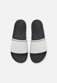 Hvide slidesandaler med en sort tekstureret sål, med en bred rem der har gentagne "Calvin Klein" logoer i sort på siden.