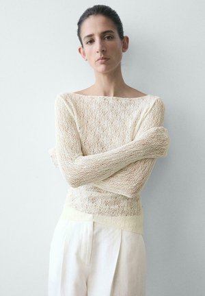 Frau mit zurückgebundenem dunklem Haar, die einen transparenten, cremefarbenen Strickpullover und lockere, helle Hosen trägt, steht mit verschränkten Armen vor einem schlichten Hintergrund.