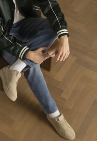 Beige suède instappers, grijze denim jeans en een groene bomberjack met witte strepen, op een houten visgraatvloer.