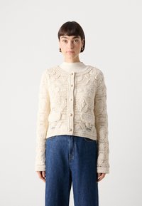 Häkelcardigan in Off-White mit floralem Muster, runder Halsauschnitt, Knopfverschluss vorne und langen Ärmeln, kombiniert mit blauen Jeans.