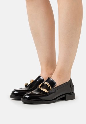 Mocasines de charol negro con un acento en tono dorado. Tacón plano, punta redondeada y textura suave. Diseño minimalista, adecuado para diversas ocasiones.