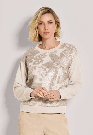 Frau mit kurzen blonden Haaren, die ein beiges Sweatshirt mit einem großen floralen Paillettenmuster und hellbeige Hose trägt, steht vor einer schlichten Wand.