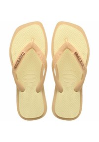 Tongs beiges avec semelles jaunes texturées, dotées d'une sangle en V et d'un détail de logo métallique sur la sangle. Conçues pour un usage décontracté.