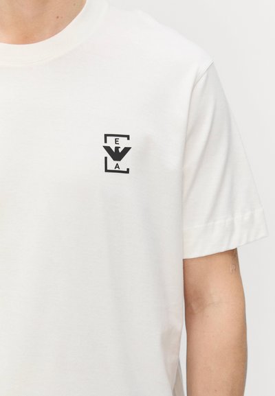 Balta medvilninė marškinė su mažais juodais logotipais kairėje krūtinės pusėje. Apvalaus kaklo dizainas, trumpomis rankovėmis ir lygia tekstūra.